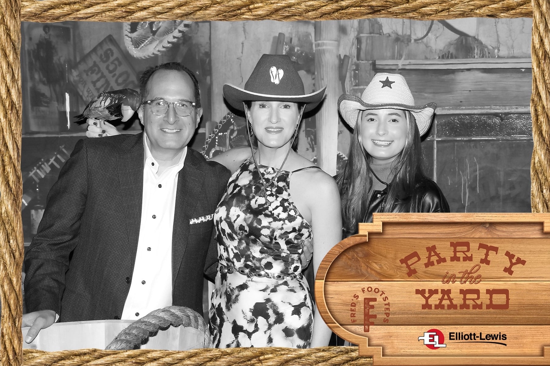 Old Time Photo Experience 1 Three people dressed in western attire smile at the camera. A rustic wooden and rope-themed frame surrounds them with a “Party in the Yard” sign and the Elliott-Lewis logo in the bottom right corner.