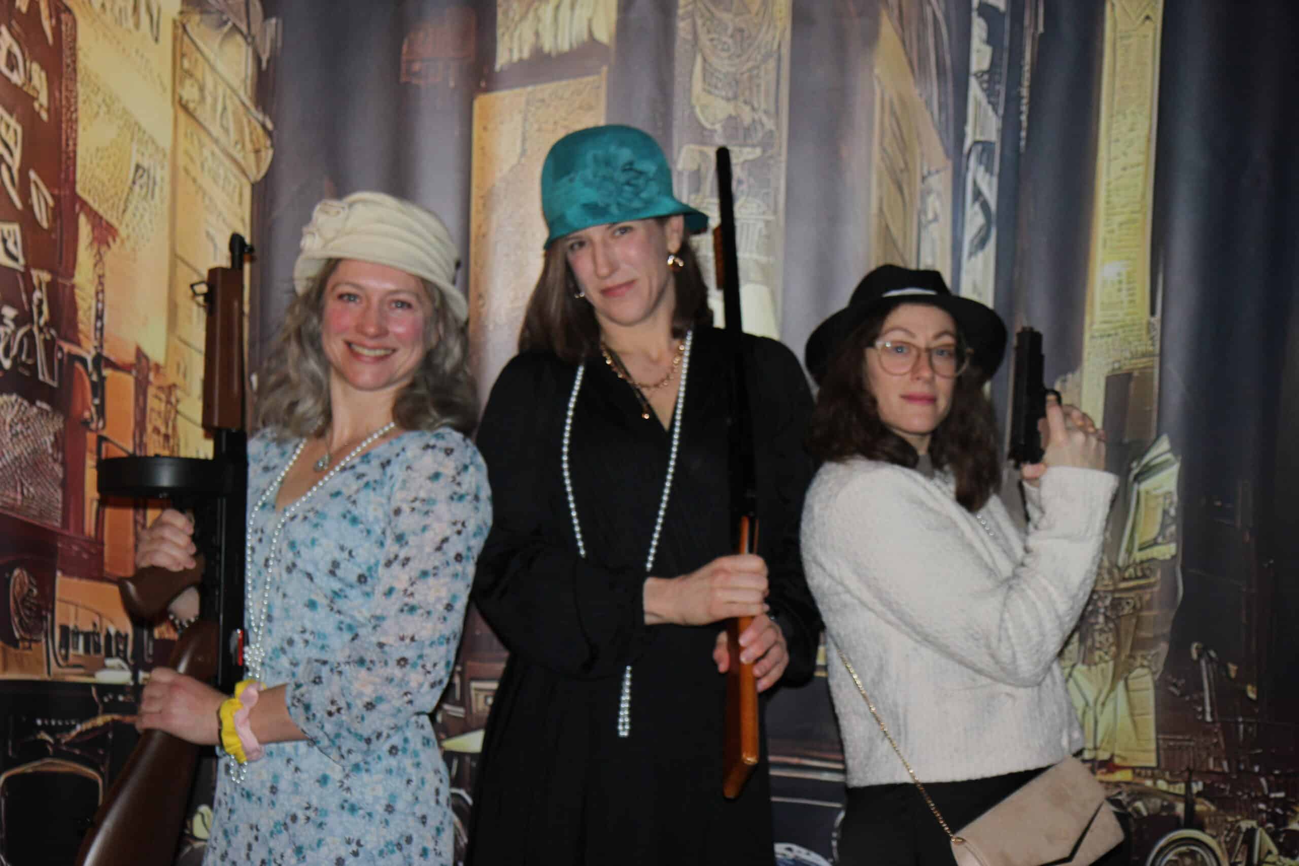 Old Time Photo Experience 5 Three women dressed in vintage 1920s-style outfits pose in front of a cityscape backdrop, each holding a toy gun and smiling.