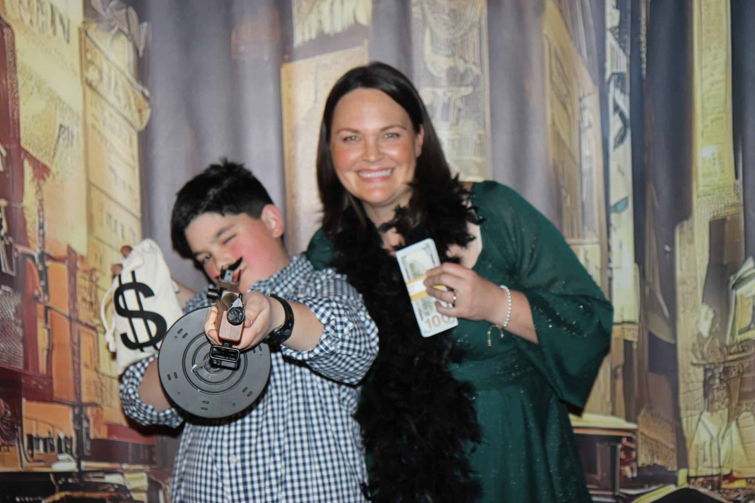 Old Time Photo Experience 2 A smiling woman in a green dress and a laughing boy with a toy gun pose together; both hold fake money props in front of a city-themed backdrop.