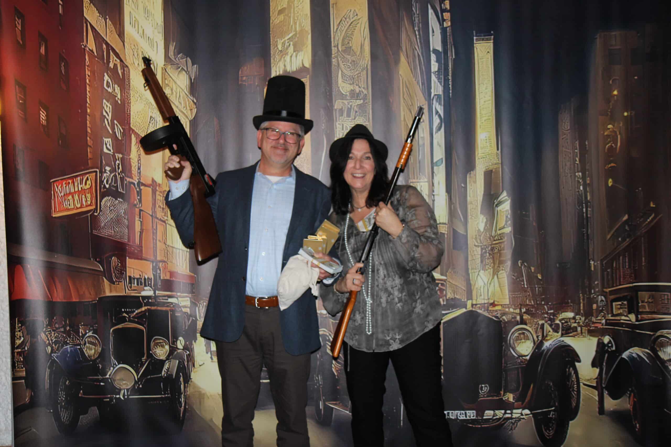 Old Time Photo Experience 9 A man and woman in vintage costumes pose with toy guns in front of a 1920s city backdrop featuring classic cars and old buildings.