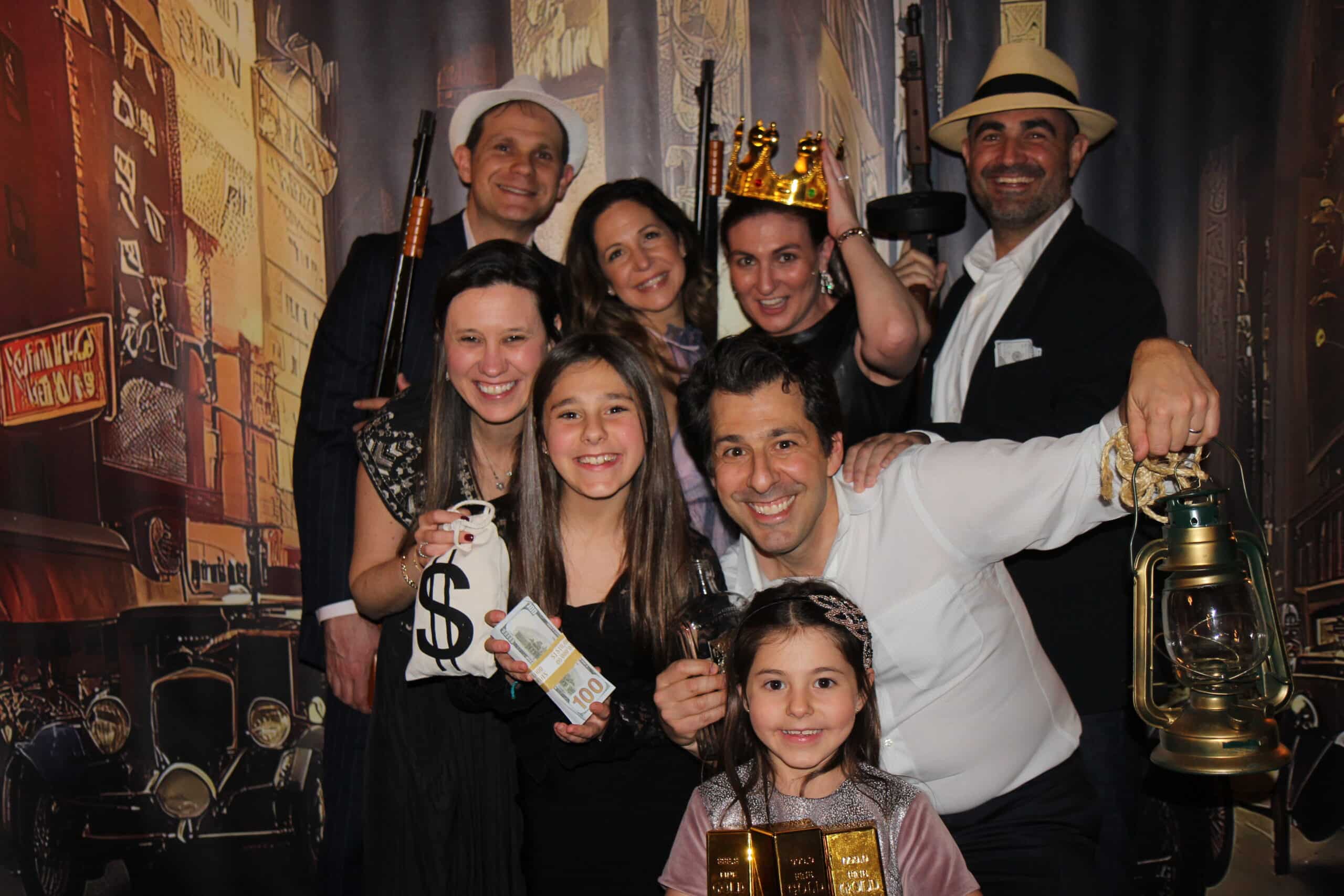 Old Time Photo Experience 10 Eight smiling people in 1920s-themed costumes hold props like gold bars, a money bag, cash, a lantern, and a crown, posing together in front of a vintage city backdrop.