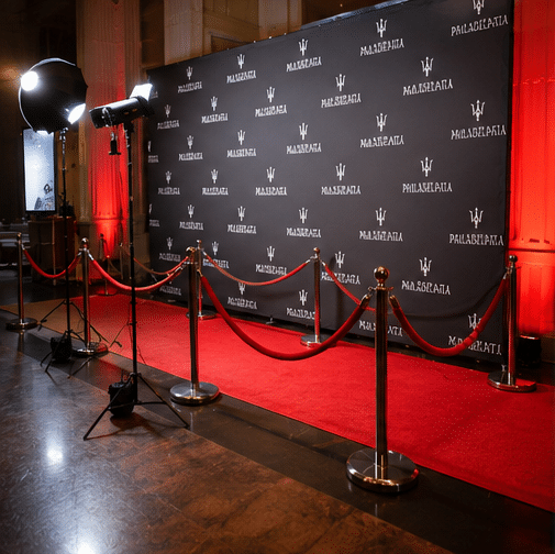 Red Carpet Photo Experience in Philadelphia 3 A red carpet event setup with stanchions, red ropes, bright lights, and a backdrop featuring Maserati logos in a grand, warmly-lit venue.