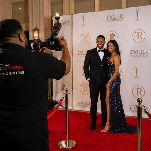 Red Carpet Photo Experience in Philadelphia 1 A couple poses and smiles on a red carpet, dressed in formal evening attire, while a photographer takes their picture in front of a step-and-repeat backdrop with Tyler Foundation and gold logos.