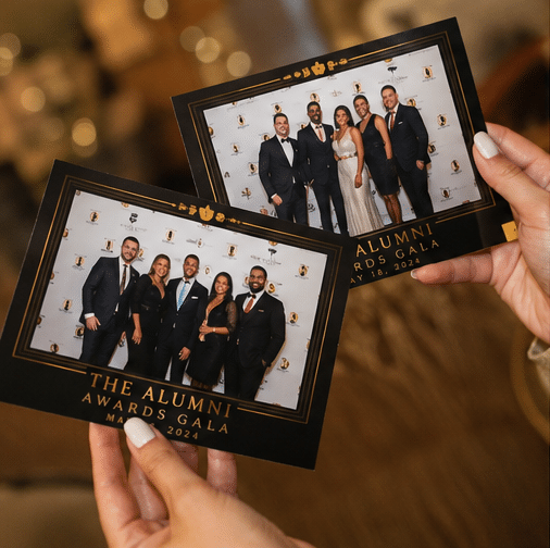 Red Carpet Photo Experience in Philadelphia 4 Two hands hold photo prints from the Alumni Awards Gala, showing groups of people in formal attire standing together and smiling for the camera.