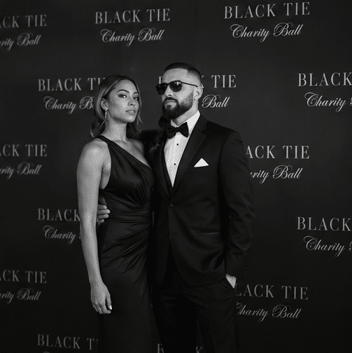 Red Carpet Photo Experience in Philadelphia 2 A woman in a black dress and a man in a black tuxedo and sunglasses pose together at a Black Tie Charity Ball event backdrop.