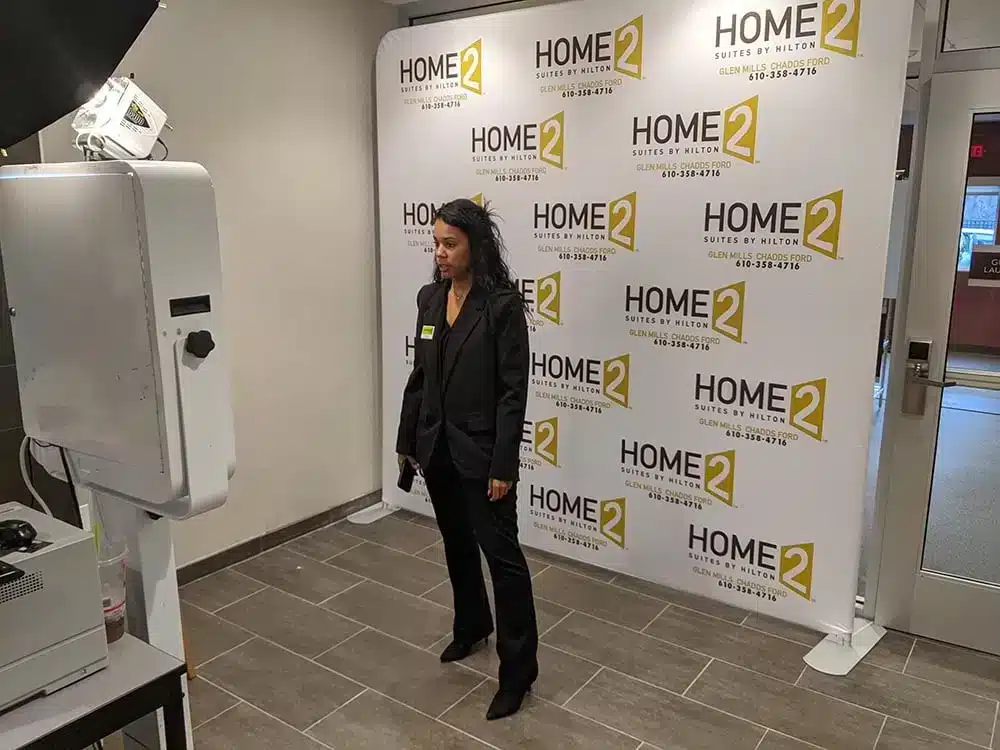 A woman in a black suit stands in front of a Home2 Suites by Hilton backdrop, facing a photo booth setup in a lobby area.